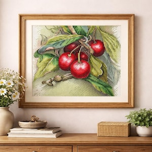 May include: A framed cross-stitch artwork depicting a bunch of red cherries with green leaves. The artwork is set against a light green background and framed in a wooden frame. The image is displayed on a wooden table.