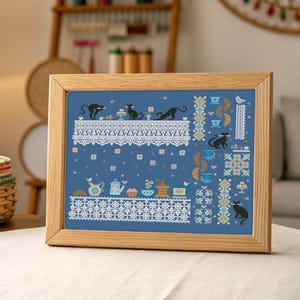 May include: A framed cross-stitch picture with a blue backdrop, white lace patterns, and black cat figures. The design incorporates teacups, pastries, and floral details, all within a light wood frame, ideal for home decor.