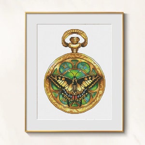 May include: A framed cross-stitch artwork featuring a gold pocket watch design. The watch face showcases a butterfly with green and yellow wings, surrounded by green and gold accents. The frame is gold.
