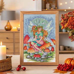 Könnte beinhalten: Ein gerahmtes Kreuzstich-Kunstwerk mit einer Fee mit roten Haaren und Flügeln, die auf einem Pilz sitzt. Die Fee trägt ein rosa-grünes Kleid. Der Hintergrund ist ein blau-grünes Blumenmuster. Der Rahmen ist aus hellem Holz.