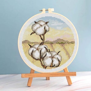 May include: A cross-stitch embroidery of cotton plants in a wooden hoop. The artwork features three fluffy white cotton bolls on brown stems against a yellow and blue background, displayed on a small wooden easel.