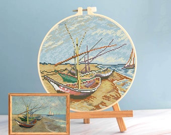 Pode incluir: Uma bordado de ponto cruz de barcos numa praia, exibido num aro de madeira sobre um pequeno cavalete. Uma pintura emoldurada da mesma cena está à frente. O bordado apresenta uma representação colorida de barcos, areia e céu.