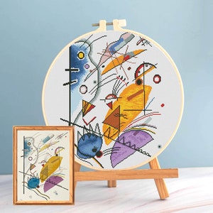 May include: A completed cross-stitch embroidery of an abstract design in a wooden hoop, displayed on a small wooden easel. The design features geometric shapes in yellow, blue, purple, and red. A framed print of the same design is also visible.