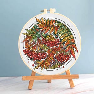 May include: A cross-stitch embroidery hoop featuring a colorful autumn design. The artwork depicts red berries and fall foliage in shades of orange, yellow, and green. The embroidery is set in a wooden hoop and displayed on a small wooden easel.