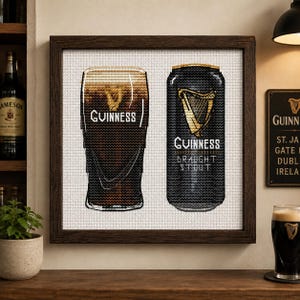 Puede incluir: Un bordado en punto de cruz enmarcado que muestra un vaso de pinta de Guinness marrón oscuro y una lata de Guinness Draught Stout negra, ambos con el logotipo del arpa dorada y texto blanco sobre un fondo de cuadrícula blanca en un marco de madera oscura.