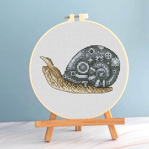 Può includere: Ricamo a punto croce di una lumaca con un design steampunk. Il guscio della lumaca è grigio con dettagli di ingranaggi e il corpo è marrone. Il ricamo è inserito in un telaio di legno ed esposto su un piccolo cavalletto di legno.