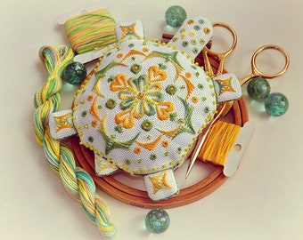 May include: A white, cross-stitched turtle-shaped pincushion with yellow and green embroidery, surrounded by embroidery floss, scissors, and glass beads. The pincushion features a detailed floral design and is set on a wooden embroidery hoop.