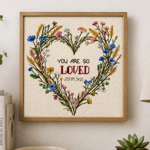 Könnte beinhalten: Ein quadratisch gerahmtes Kreuzstich-Stickbild mit einem herzförmigen Kranz aus bunten Wildblumen in Blau, Gelb, Rosa und Grün. Der mittlere Text lautet "YOU ARE SO LOVED" in Schwarz und Rot, gefolgt von "JOHN 5:16" in schwarzer Schrift.
