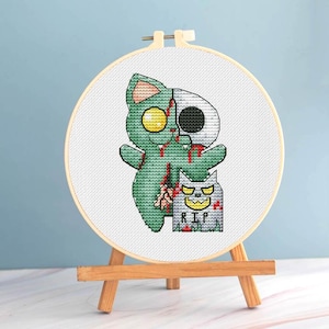 May include: A cross-stitch embroidery hoop featuring a green zombie cat and a half-skull cat design. The artwork includes a tombstone with "RIP" and a small wooden easel. The background is a light blue.