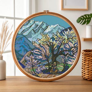 May include: A cross-stitch landscape scene in a wooden embroidery hoop. The artwork depicts a mountain range in shades of blue and green, with a tree in the foreground. The tree has yellow and white leaves. The hoop is displayed on a wooden table.