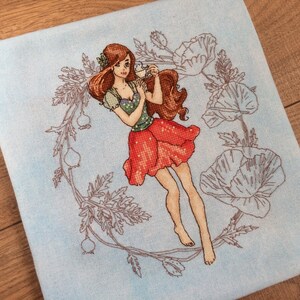 Könnte beinhalten: Kreuzstich-Kunstwerk mit einer Frau mit langen, rotbraunen Haaren, die ein grünes und rotes Kleid trägt und eine Teetasse hält. Sie ist von einem Blumenkranz und Mohnblumen umgeben, alles auf hellblauem Hintergrund. Das Kunstwerk ist detailliert und farbenfroh und zeigt ein skurriles Thema.