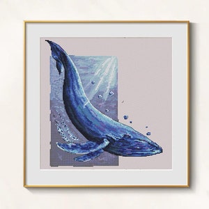 May include: A framed cross-stitch art piece featuring a blue whale swimming underwater. The whale is depicted in shades of blue with bubbles and light rays. The artwork is framed in a gold-colored frame.