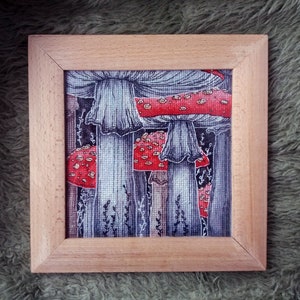 Könnte beinhalten: Gerahmte Illustration von roten Pilzen mit weißen Punkten und grauen Kappen, vor einem Hintergrund aus grauem und schwarzem Laub. Das Kunstwerk wird in einem hellen Holzrahmen präsentiert. Detaillierte, handgezeichnete Darstellung.