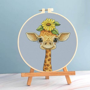 May include: A cross-stitch embroidery of a giraffe wearing a sunflower crown, displayed in a wooden hoop. The giraffe has a tan and brown pattern, with large, expressive eyes. The background is a light blue fabric, and the hoop is mounted on a small wooden easel.
