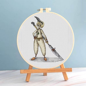 May include: A cross-stitch embroidery of a cartoon character in a wooden hoop. The character is wearing a cream-colored outfit, a horned helmet, and holding a large sword. The embroidery is displayed on a small wooden easel against a light blue background.