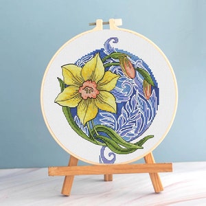 May include: A completed cross-stitch embroidery of a yellow daffodil with green leaves and a pink center, set against a blue background. The design is framed in a wooden hoop and displayed on a small wooden easel.