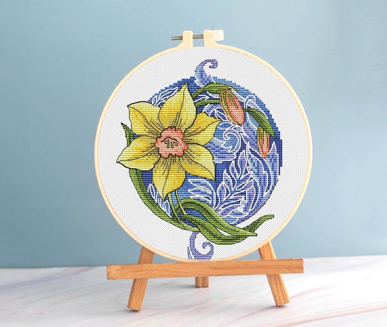 May include: A completed cross-stitch embroidery of a yellow daffodil with green leaves and a pink center, set against a blue background. The design is framed in a wooden hoop and displayed on a small wooden easel.