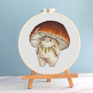 Könnte beinhalten: Ein Kreuzstich-Stickrahmen mit einer Cartoon-Pilzfigur mit einem wütenden Gesichtsausdruck. Der Pilz hat einen beigen Körper, eine große beige Kappe und eine Halskette aus gelben Blättern. Der Hintergrund ist ein Farbverlauf aus Orange und Braun.