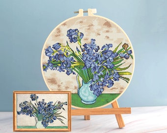 Pode incluir: Uma bordado em ponto cruz de íris azuis num vaso turquesa, exibido num bastidor de madeira. Uma impressão emoldurada menor do mesmo arranjo floral também é visível. A obra de arte está definida contra um fundo azul claro.