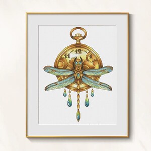 May include: Cross-stitch art of a golden pocket watch with a dragonfly design. The dragonfly has blue and green wings and is adorned with teardrop-shaped jewels. The artwork is framed in a gold-colored frame.