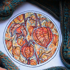 May include: A cross-stitch embroidery of orange and yellow Chinese lantern plants within a white circle. The artwork is surrounded by teal and brown knitted fabric and a skein of colorful thread.