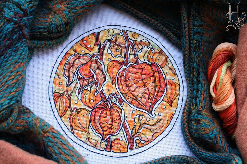 May include: A cross-stitch embroidery of orange and yellow Chinese lantern plants within a white circle. The artwork is surrounded by teal and brown knitted fabric and a skein of colorful thread.