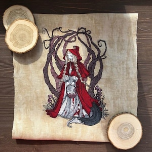 May include: Cross-stitch art featuring a girl in a red hooded cloak, with a wolf tail, standing before a dark, twisted tree. The artwork has a vintage, aged appearance. Wooden slices are placed around the artwork.