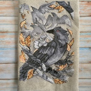 May include: Cross-stitch artwork featuring a large black crow perched on a branch, surrounded by other crows and autumn leaves. The artwork is on a beige fabric background, with a wooden plank backdrop.