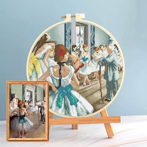 May include: A cross-stitch embroidery of a Degas painting, featuring ballerinas in a dance studio. The artwork is displayed in a wooden hoop and a framed print, both on a wooden easel. The scene is rendered in shades of blue, white, and brown.