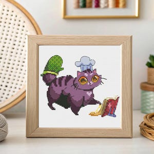 May include: Cross-stitch art of a purple cat wearing a chef's hat and oven mitt, with a cookbook and spilled food. The framed artwork has a light wood frame and is displayed on a white surface.