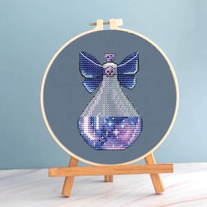 May include: A cross-stitch design of a potion bottle with a skull-shaped stopper and a bow. The bottle is filled with a galaxy-like pattern of purple, blue, and white. The design is set in a wooden embroidery hoop and displayed on a small wooden easel.