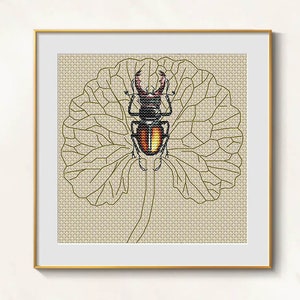 Puede incluir: Arte de punto de cruz enmarcado, con un escarabajo detallado con detalles rojos y amarillos sobre una hoja beige. La obra, en un marco dorado, resalta la intrincada costura y el tema natural.