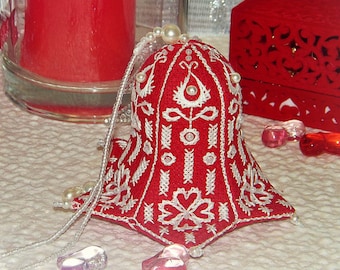 Op de afbeelding: Een rode, klokvormige ornament met ingewikkelde witte borduursels en parelaccenten. Het ornament heeft een kralenketting om op te hangen. De achtergrond bevat een rode kaars en een decoratieve doos, wat wijst op een feestelijk thema.