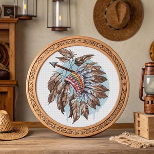 Peut inclure: Une broderie au point de croix ronde encadrée représentant une coiffe amérindienne avec des plumes marron, rouges et jaunes. Une flèche est placée en travers de la coiffe. Le cadre est en bois brun clair.