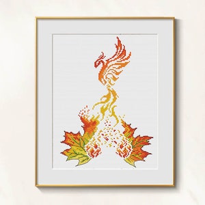 May include: Framed cross-stitch art featuring a phoenix rising from flames and maple leaves. The phoenix is rendered in shades of orange and yellow, with the leaves in green, orange, and yellow. The artwork is framed in a gold-colored frame.