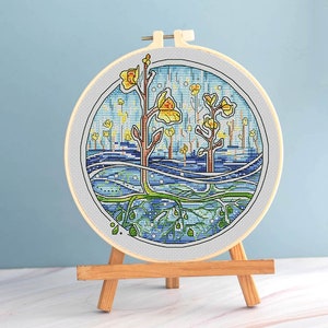 Puede incluir: Un bastidor de bordado de punto de cruz con un paisaje colorido con &aacute;rboles y ra&iacute;ces estilizados. La obra de arte utiliza tonos de azul, amarillo y verde, creando una escena caprichosa inspirada en la naturaleza. El bordado se exhibe en un peque&ntilde;o caballete de madera.