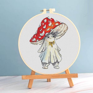Könnte beinhalten: Kreuzstich-Stickerei eines Pilzcharakters mit roter und weiß gepunkteter Kappe, der ein weißes Outfit und eine goldene Halskette trägt. Die Stickerei befindet sich in einem Holzreifen, der auf einer kleinen Holzstaffelei vor hellblauem Hintergrund ausgestellt ist.
