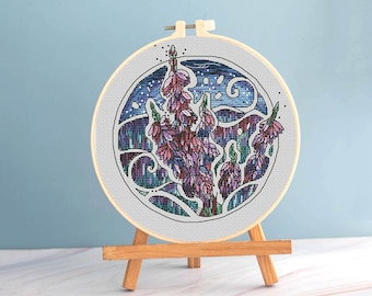 Puede incluir: Un bordado de punto de cruz de flores moradas y rosas dentro de un círculo con borde blanco. El diseño está sobre un fondo azul, montado en un bastidor de bordado de madera y expuesto en un pequeño caballete de madera.
