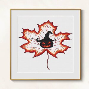 Puede incluir: Un diseño de punto de cruz enmarcado con un tema de Halloween de hoja de arce. La hoja es naranja y roja, con una calabaza negra con un sombrero de bruja en el centro. Una pequeña araña cuelga de un hilo.