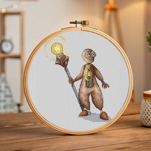 May include: Cross-stitch artwork featuring a quirky character holding a lightbulb. The character is brown with goggles and a zipper design. The lightbulb is yellow, and the artwork is framed in a wooden hoop.