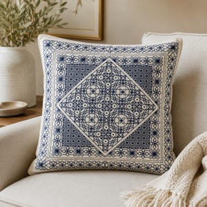 Könnte beinhalten: Ein quadratisches Zierkissen mit einem cremefarbenen Stoffbezug und aufwendiger dunkelblauer Stickerei. Das Design besteht aus einem zentralen Rautenmuster, das von geometrischen Rändern und blumenartigen Motiven umgeben ist.