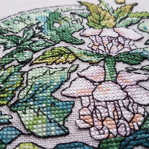 May include: Close-up of a cross-stitch embroidery featuring a floral design. The artwork showcases a central flower in shades of pink and peach, surrounded by green leaves and foliage. The stitches are detailed and the background is a light neutral color.