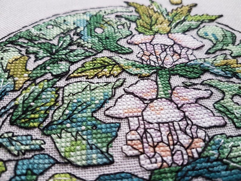May include: Close-up of a cross-stitch embroidery featuring a floral design. The artwork showcases a central flower in shades of pink and peach, surrounded by green leaves and foliage. The stitches are detailed and the background is a light neutral color.