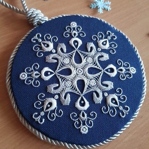May include: A round, navy blue embroidered ornament with a white snowflake design. The ornament has a silver rope border and a silver hanging loop. Decorative snowflakes are scattered around the ornament.