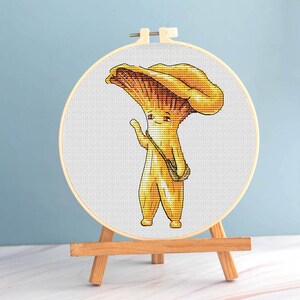 Könnte beinhalten: Ein Kreuzstich-Stickrahmen mit einer Cartoon-Pfifferling-Pilzfigur mit einer Umhängetasche. Der Pilz ist gelb und orange, mit einem freundlichen Gesichtsausdruck. Die Stickerei wird auf einer kleinen Holzstaffelei präsentiert.