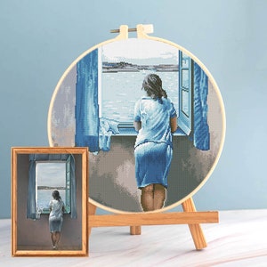 May include: A cross-stitch embroidery hoop and a framed painting depict a woman gazing out a window at a seascape. The embroidery features a detailed pixelated image in shades of blue. The painting is in a wooden frame.