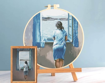 Pode incluir: Um bastidor de bordado em ponto cruz e uma pintura emoldurada retratam uma mulher olhando por uma janela para uma paisagem marinha. O bordado é uma imagem pixelizada detalhada em tons de azul. A pintura está em uma moldura de madeira.