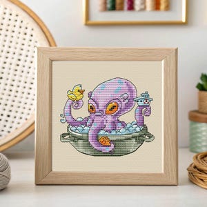 May include: A framed cross-stitch depicts a purple octopus in a bathtub with a yellow rubber ducky and a lighthouse. The octopus has orange eyes and blue spots. The artwork is set against a neutral background, framed in light wood.