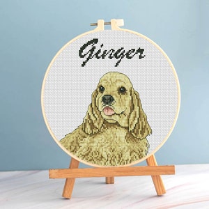 May include: A cross-stitch embroidery hoop featuring a golden Cocker Spaniel with the name "Ginger" above. The dog's portrait is detailed, with a light brown and tan color palette. The hoop is displayed on a small wooden easel.