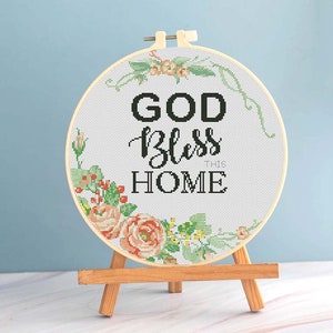 May include: A cross-stitch embroidery hoop with the words "GOD Bless THIS HOME" in black. The design is surrounded by a floral wreath in shades of peach, green, and yellow. The hoop is displayed on a small wooden easel.